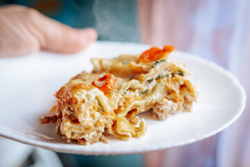 A close-up of a freshly baked slice of lasagna with melted cheese, cherry tomatoes, and herbs on a white plate. The dish appears hot and steaming, ready to be enjoyed.