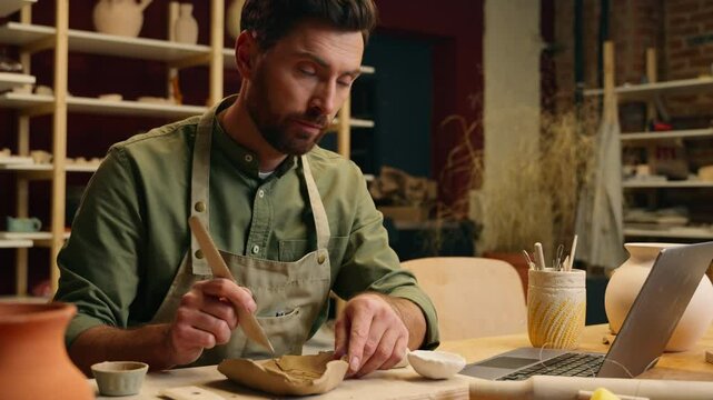 Caucasian male man thoughtful creative small business owner businessman guy potter craftsman making stoneware tool watching laptop video tutorial pottery lesson online computer device indoors studio