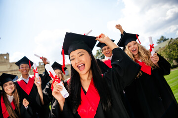 Smiling young graduates celebrating on campus in mortarboards and gowns on a sunny summer day after...