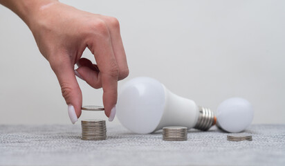 Female hand stacking coins in stacks, next to LED lights, symbolizing energy saving and efficiency. LED light bulb is modern technology, while money highlight is smart energy conservation.