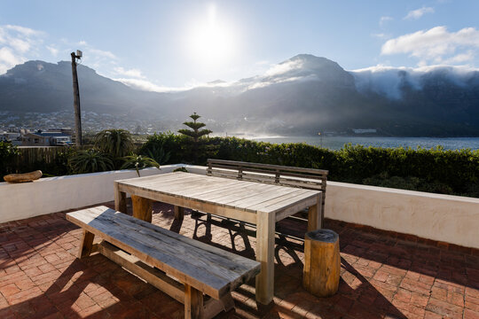 Wooden table on sunlit patio overlooking mountains and ocean, ideal for relaxing, copy space