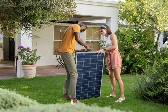 Diverse couple installing solar panel in garden, enjoying sustainable living on vacation - Powered by Adobe