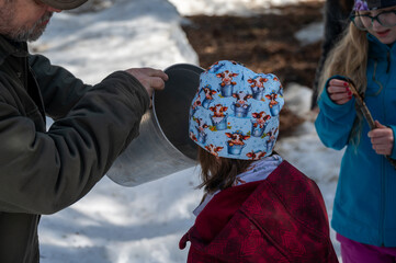 In a snowy woodland, children excitedly gather maple sugar water with a joyous spirit. Wearing...