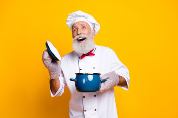 Joyful senior chef holding a cooking pot against a yellow backdrop, celebrating culinary skills and a passion for cooking