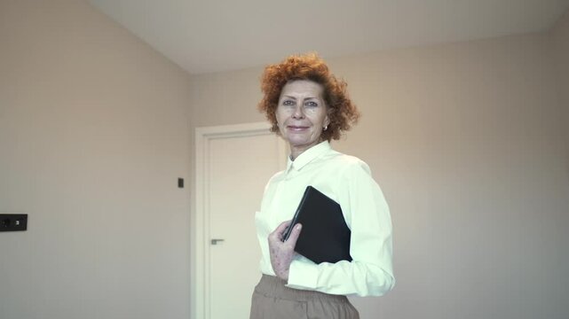 Mature female real estate agent stands in an empty apartment, holding folder with documents. She confidently looks at the camera, dressed professionally, ready to show property to potential buyers. 