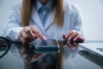 Woman doctor in white coat sitting working at desk in the clinic office room, using smartphone, close shot of woman holding hand finger over smartphone screen.