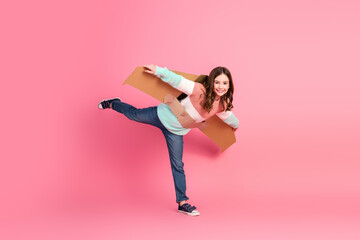 Charming girl playing pretend airplane with cardboard wings against pink background, expressing...
