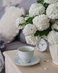 A cup of coffee, an alarm clock and a vase of white flowers on the bedside table. Tea time, coffee time