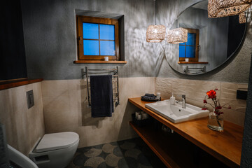 A cozy bathroom with warm lighting, a wooden countertop, a round mirror, and rustic woven lamps. A red rose in a glass vase adds elegance to the decor.