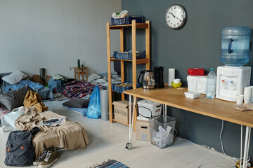 Wooden table with water cooler, disposable kitchenware, teapots, empty plastic bottle and containers standing by grey wall