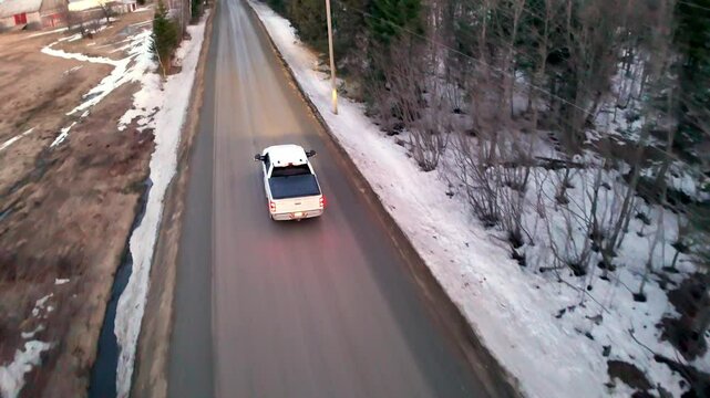 A truck travels down a gravel road surrounded by bare trees and patches of lingering snow. A small creek flows alongside, creating a serene winter atmosphere.