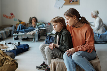 Young woman and her son in casual apparel sitting on sleeper in refugee camp and talking against...