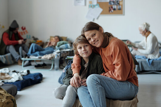 Happy young woman embracing her cute son while both sitting on sleeper and looking at camera against homeless people