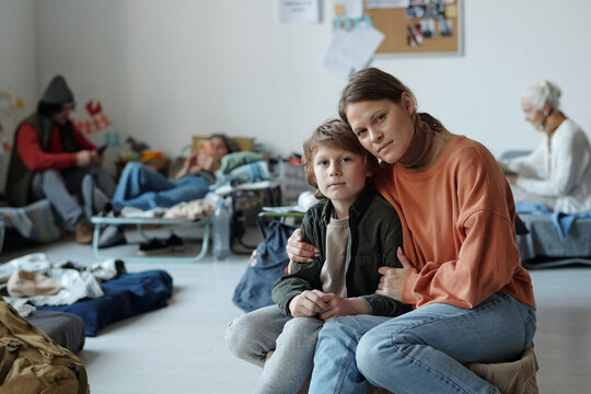 Serious woman embracing her little son while both sitting in front of camera in crisis camp against homeless people