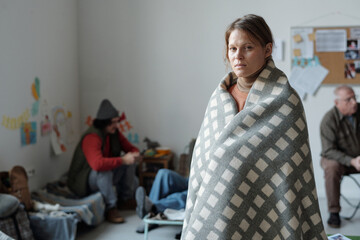 Young miserable woman wrapped in plaid or blanket looking at camera while standing in refugee camp...