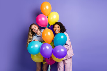 Two young women holding colorful balloons against a purple backdrop, smiling joyfully, embracing...
