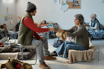 Young man passing cup of tea to mature female refugee while both sitting on their sleepers among...