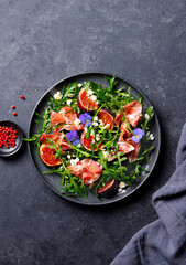 Arugula salad with fresh figs and prosciutto, jamon and edible flowers in a plate. Restaurant serving, plating. Dark background. Top view.