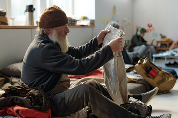 Side view of senior male refugee with long grey beard sitting on his sleeper and taking pants out of backpack in crisis camp