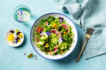 Green salad with watermelon radish and edible flowers in a bowl. Blue background. Top view.