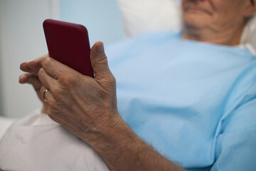 Close-up of senior man lying on bed in the ward and texting message on mobile phone