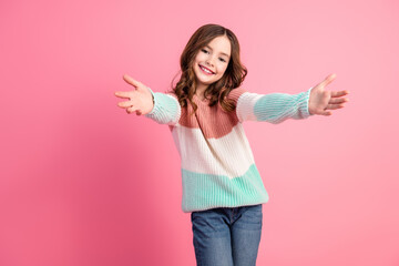 Charming pretty girl with brown hair extending arms in a welcoming gesture wearing a stylish colorful sweater against a pink background