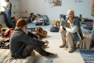 Joyful senior female refugee with book in hands laughing at joke of elderly man sitting on sleeper...