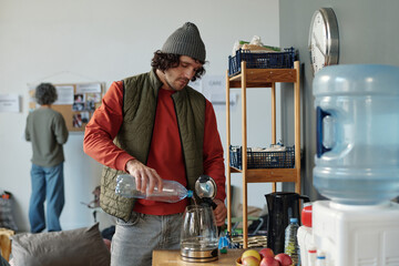 Young male refugee pouring pure water from plastic bottle in teapot while standing by table with...