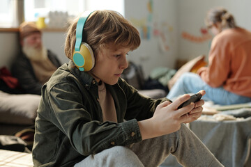 Serious boy in headphones looking at screen of mobile phone in his hands while playing video game or watching movie in camp
