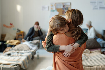 Rear view of young single mother holding her youthful son on hands while sad boy embracing her in camp for those in need