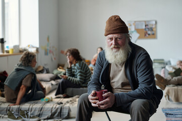 Senior man with grey beard holding mug and looking at camera while sitting in crisis camp for...