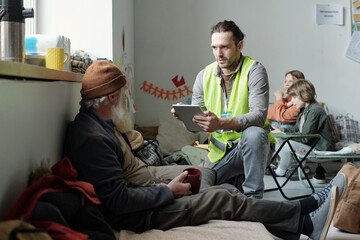 Young man with tablet entering personal data of senior male refugee in database while sitting on...