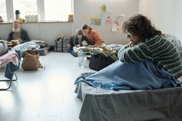 Lonely female refugee in striped sweater sitting on sleeper in crisis camp with homeless people and...