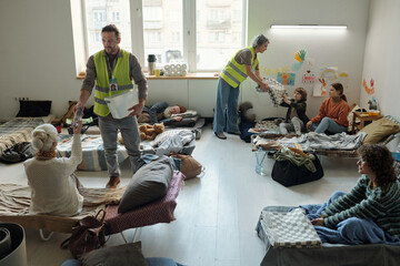 Two volunteers in fluorescent jackets passing water and blankets to homeless people sitting on...