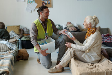 Young volunteer in casualwear and reflective jacket passing bottle of water to senior female refugee sitting on sleeper in front of him