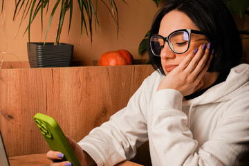 A woman wearing glasses and a white hoodie using her smartphone while working on a laptop. A cozy...