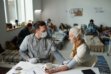 Young doctor in medical scrubs and protective mask sitting by table in front of senior female...