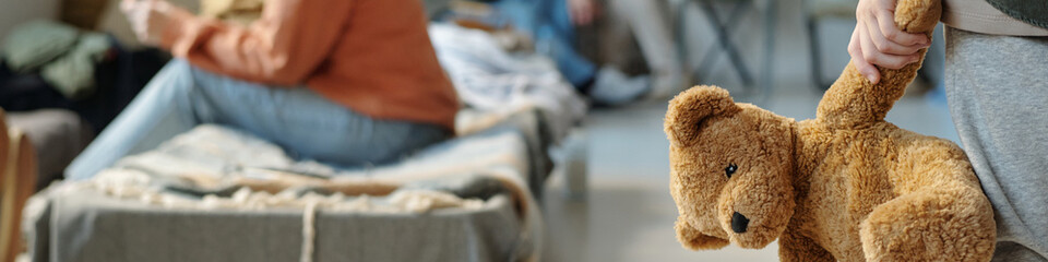Header of hand of little boy in grey sweatpants holding brown teddybear while standing in front of...