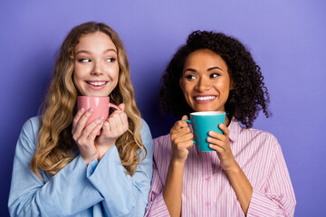 Two joyful young women in stylish pajamas enjoying warm drinks and smiling on a vibrant purple...