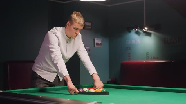 Billiards trainee in shirt and gray trousers arranges colorful billiard balls in triangle on green pool table. Overhead lights cast warm glow as he focuses with calm concentration in stylish lounge