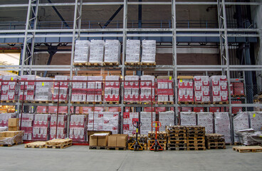 A well-organized warehouse storage area is filled with stacked pallets labeled and ready for distribution. Pallet jacks and additional empty pallets are neatly arranged on the floor below the shelves.