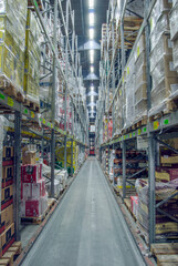 Tall shelves filled with various packaged goods line a long, narrow aisle in a large warehouse....
