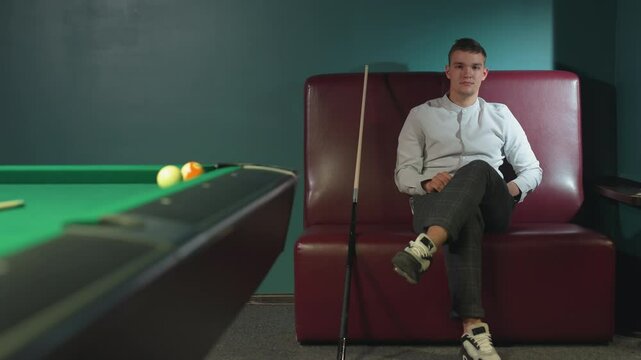 Male in white shirt and plaid pants sits on red sofa, rolling orange billiard ball in hand with serious expression. Cue stick leans beside him. Green pool table with scattered balls in background
