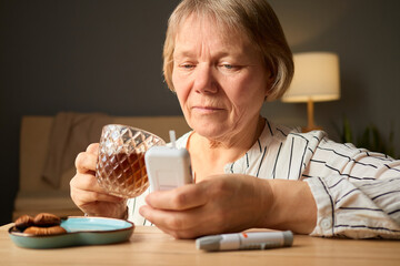 A senior woman sits comfortably at a table, carefully checking her blood sugar levels with a glucometer. She holds a cup of tea in one hand and has a plate of cookies beside her