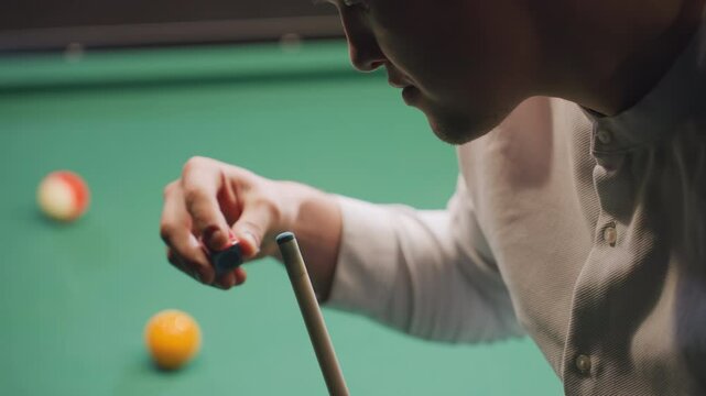 Player in white shirt chalking cue stick before taking shot on green pool table. Close-up highlights preparation, focus, and skill, with blurred billiard balls in background