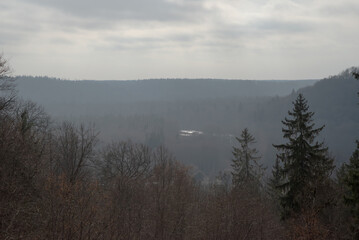 A misty valley landscape with layers of hills, trees, and a cloudy sky. The scene has a moody, overcast atmosphere with muted colors.