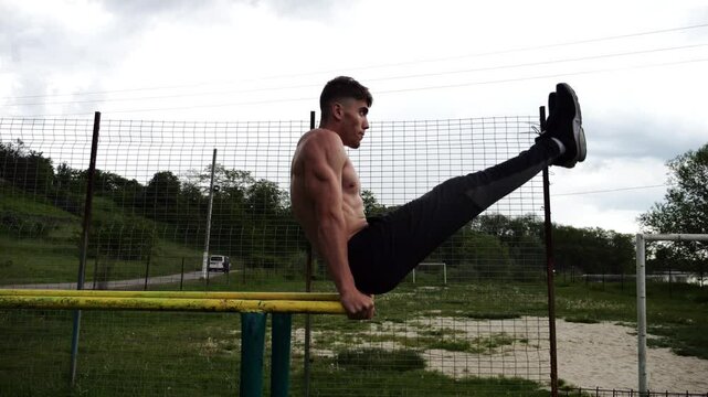 Strong man trains abdominal muscles raising legs on parallel bars at sports ground. Young shirtless sportsman exercising at nature. Sporty athlete working out at nature. Concept of sportive lifestyle
