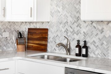 A kitchen faucet detail with white cabinets, decorations on a white marble countertop, and a marble herringbone tile backsplash.