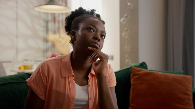 Pondering pensive thoughtful African American ethnic woman puzzled female model lady homeowner client thinking touching chin deep thoughts solving problem solution sitting couch indoors at home room