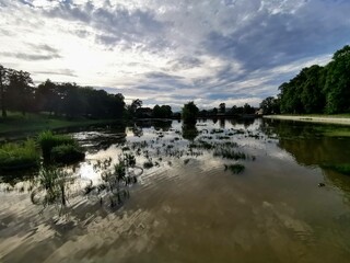 Peaceful pond in Kudowa-Zdrój, Poland, with reflective waters, a small island, and a dramatic sky, framed by verdant trees